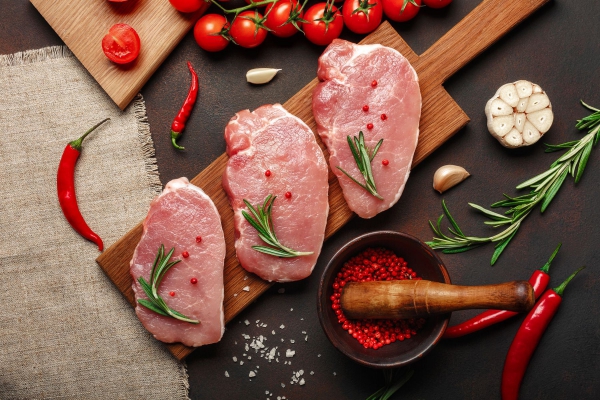 Pieces of raw pork steak on cutting board with cherry tomatoes, rosemary, garlic, pepper, salt and spice mortar on rusty brown background